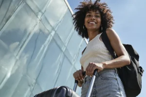 Jovem sorridente com mochila nas costas e segurando a alça de uma mala de viagem, pronta para o intercâmbio no exterior após organizar o seu Apostilamento de Haia.