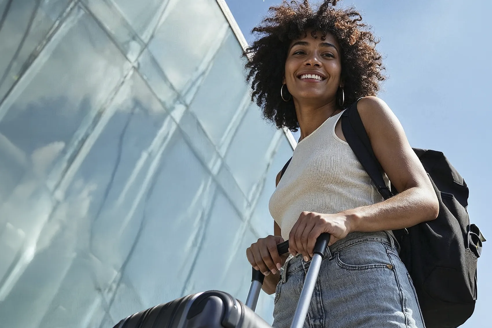 Jovem sorridente com mochila nas costas e segurando a alça de uma mala de viagem, pronta para o intercâmbio no exterior após organizar o seu Apostilamento de Haia.